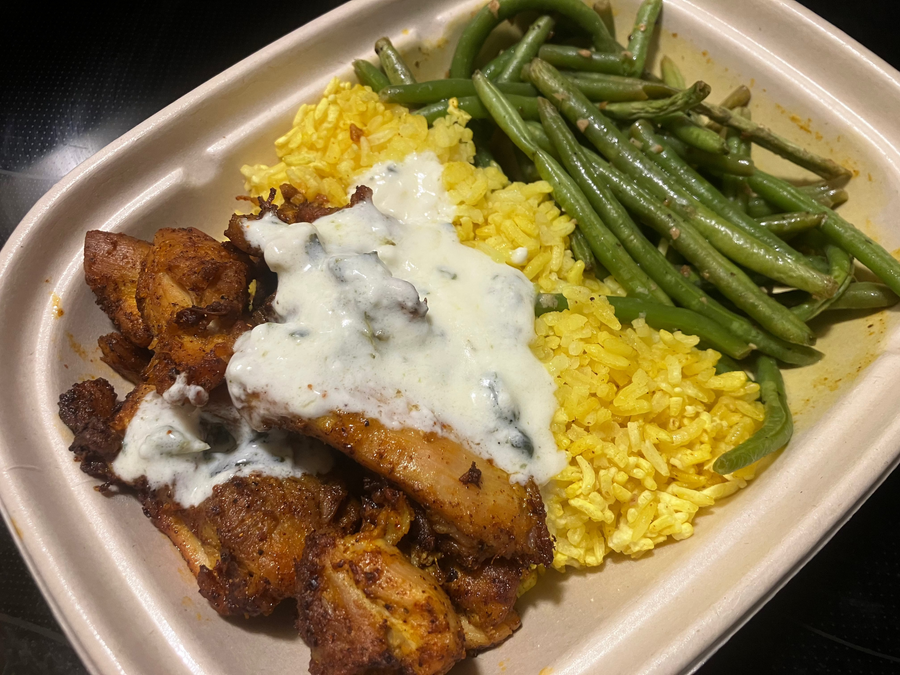 A close-up of a Shawarma Chciken Means meal in a compostable container with chicken, yellow rice, green beans, and a white creamy sauce.