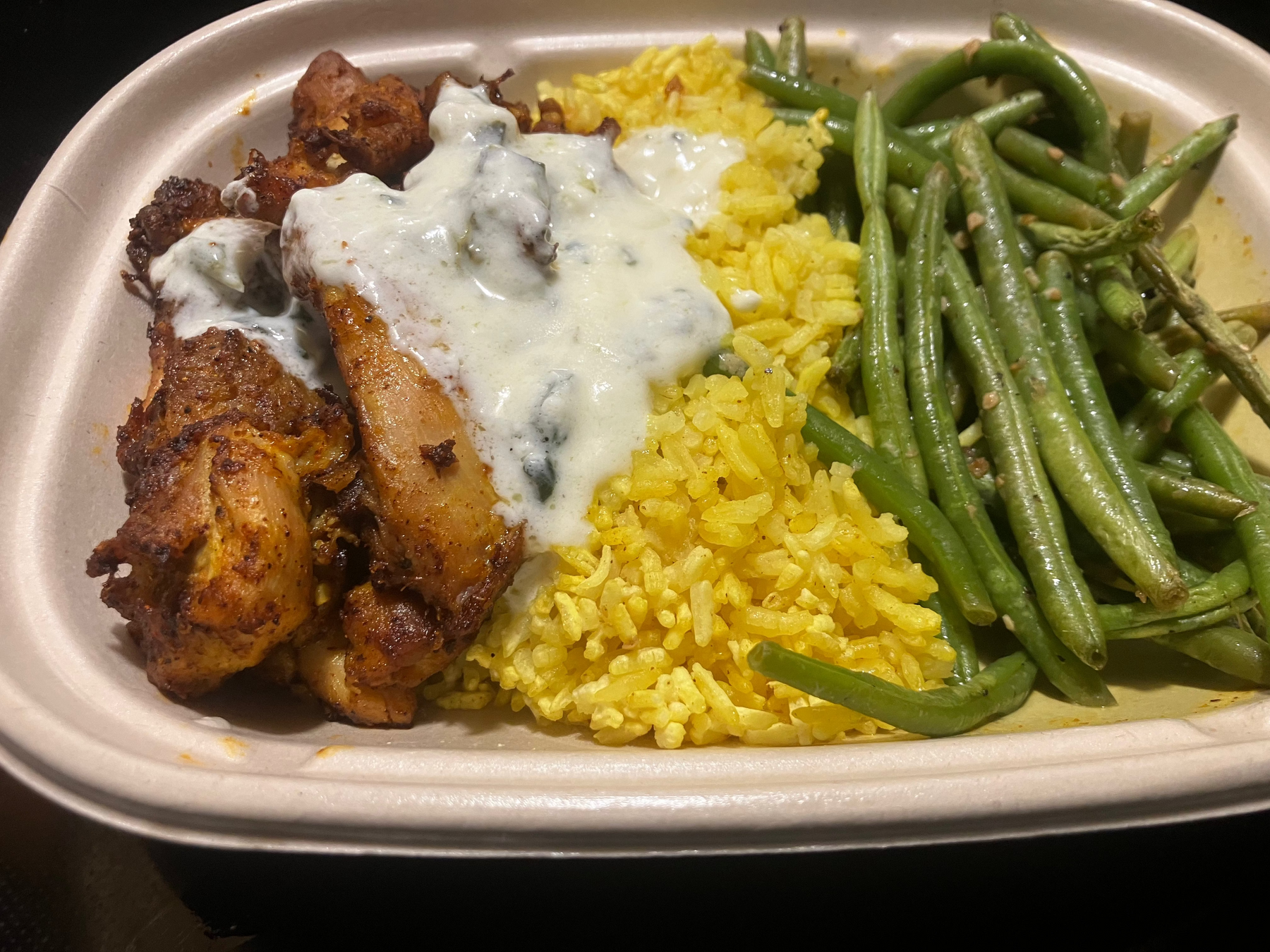 A close-up overhead shot of a prepared Shawarma Chicken in a compostable container, featuring yellow rice, green beans, and chicken topped with a white creamy sauce.